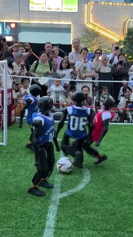 A group of black humanoid robots wearing blue and red soccer jerseys are playing soccer on a green artificial turf field. The robots have black heads with red eyes and black bodies. Some of the robots are wearing blue jerseys with the number 06, and others are wearing red jerseys. One robot in a blue jersey is kicking the soccer ball. A crowd of people are standing behind a red and white rope, watching the robots play. The people are of various ages and ethnicities, and many are holding up their phones to take pictures or videos. The background shows a building with lights and signs. The scene appears to be an outdoor event or exhibition.