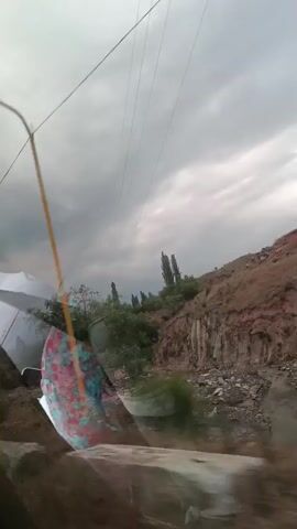 The video shows a view from inside a vehicle, looking out through the windshield. The sky is cloudy, with dark clouds suggesting an approaching storm. A rural landscape is visible outside, featuring hills, sparse vegetation, and a few tall trees. There is also evidence of some sort of construction or debris on the side of the road. A blurry reflection of what appears to be a person in a colorful shirt can be seen on the windshield. The vehicle is moving, as indicated by the slight motion blur in the footage. The overall atmosphere is somewhat somber due to the weather.