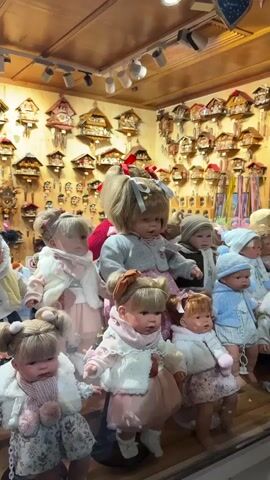 A collection of lifelike baby dolls are displayed in a store window. The dolls are dressed in various outfits, many with pink and white accents. In the background, numerous cuckoo clocks hang on the wall. The scene is well-lit, highlighting the details of the dolls and the clocks.