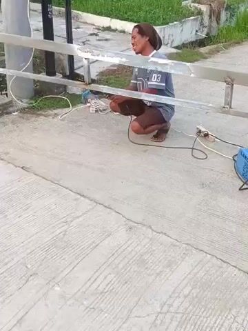 A man wearing a brown headwrap, a grey t-shirt with the number "03" on it, and brown shorts is crouching next to a metal fence. He is holding an angle grinder in his right hand and appears to be grinding the fence. A welding machine and some cables are on the ground next to him. The man then stands up, holding the angle grinder. He is on a concrete surface, with grass and some buildings in the background.