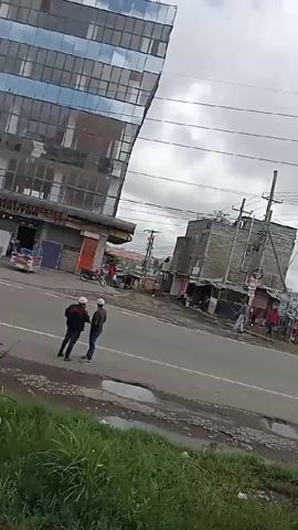 The video shows a street scene in what appears to be a developing country. A modern glass building stands on the left side of the frame, juxtaposed against more modest structures across the street. Two men are standing on the side of the road, appearing to be engaged in conversation. Across the street are small shops or stalls, with a few individuals walking or standing nearby. The sky is overcast, and power lines crisscross overhead. There is some green vegetation in the foreground, along with puddles or standing water on the road surface. The overall impression is of a place where modern development coexists with simpler, more traditional ways of life.