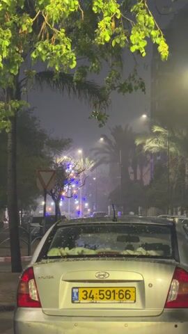 The camera is placed behind a parked silver Hyundai car, showing a foggy street scene at night. The car's license plate, 34-591-66, is visible. Trees line the street, some adorned with strings of blue and other colored lights. Streetlights illuminate the scene, adding to the fog. A yield sign is also visible on the left side of the street.