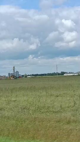 The video shows a rural landscape with a field of tall green grass in the foreground and middle ground. In the distance, there are several agricultural buildings, including silos and what appear to be storage or processing facilities. The sky is filled with dramatic, puffy clouds, suggesting a dynamic weather pattern, possibly a partly cloudy or overcast day. The lighting appears to be soft, indicating it could be morning or late afternoon. The overall impression is one of agricultural activity in a relatively undeveloped area. No people are visible. The scene is set in Shumilina, Belarus, a region known for its agricultural land. The camera appears to be moving, likely from a vehicle, as indicated by the slight motion blur and the changing perspective of the landscape.