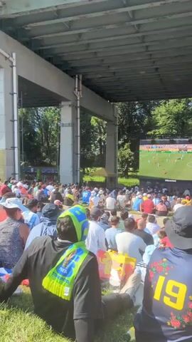 The video shows a large crowd of people sitting on a grassy area under a bridge. They are facing a very large outdoor screen where a soccer match is being broadcast. The screen is positioned in front of trees and a blue sky is visible above the trees. The bridge structure is made of concrete pillars and a gray ceiling. People are sitting on blankets and the grass, many are wearing light-colored clothing. Some people are wearing soccer jerseys. The focus is on the crowd watching the screen, with the screen itself appearing as a bright rectangle in the distance. The sun is shining, casting shadows. In the foreground, the back of a person wearing a dark blue shirt with the number 19 and a floral design is visible. To the left, another person wears a black jacket with a bright yellow and green scarf. The atmosphere appears to be relaxed, with people gathered to watch the sporting event together.