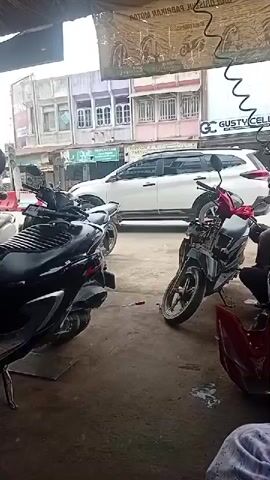 The video shows a street scene viewed from inside a covered area. In the foreground, to the left, is a black scooter. To the right, a partially disassembled motorcycle is visible, with a red plastic part on the ground in front of it. Further back, a white SUV is driving away from the camera. Beyond the SUV, there are buildings with multiple windows and balconies. A sign for "GUSTYCELL" is visible on one of the buildings. A man in a light-colored shirt and dark shorts walks from right to left in the middle ground, passing behind the motorcycles. The lighting suggests it is daytime, possibly overcast.
