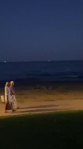The video shows a nighttime scene at a beach. The sky is dark blue, and the sea is dark blue with some light reflecting on the water. There is a sandy beach with some grass in the foreground. In the distance, there are a few lights visible on the water, possibly from boats. In the foreground, there are two people walking on a path near the beach. They are both wearing light-colored clothing. There is a lamppost with a light on it, and a trash can next to it. The moon is visible in the sky, and its reflection is on the water.