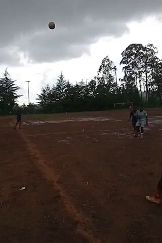 The video captures a soccer game on a muddy field. Several young soccer players wearing different colored jerseys are visible; one wears a grey jersey with the number 5, another a navy jersey with the number 13. The ball is in the air at the beginning, and later, the players are shown running and chasing the ball. The field appears waterlogged. In the background, there are tall trees and some buildings under a cloudy sky.