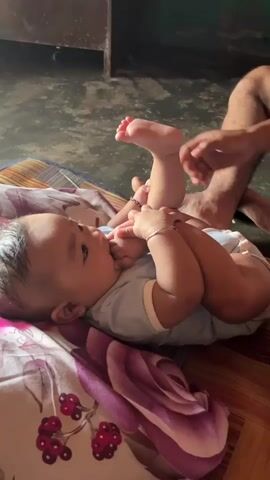 A baby lies on their back on a patterned blanket. The baby is wearing a light blue onesie with white trim. The baby is holding their feet and putting their toes in their mouth. The baby has dark hair and is wearing a gold earring and a silver bracelet. In the background, a person's legs and hands are visible, suggesting they are interacting with the baby. The environment appears to be indoors, with a concrete floor visible. The lighting suggests it is daytime.