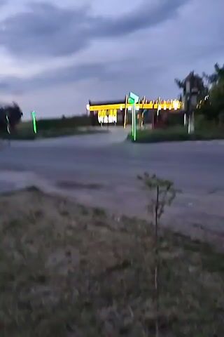 The video shows a car wash at dusk. The sky is cloudy and a pale gray. The car wash has bright green lights on its poles and on the entrance. The words "POLISH WASH" are lit up in yellow letters on the entrance. There is a paved road in front of the car wash, and grass and weeds grow on the side of the road. A small tree is growing on the right side of the frame. The video is shaky and seems to be taken from a moving vehicle.