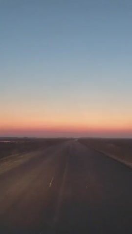 A video shows a straight asphalt road stretching into the distance. The sky above the horizon is a gradient of colors, starting with a pale blue at the top, transitioning to a soft pink and orange band just above the horizon, and then a darker shade where it meets the land. The landscape on either side of the road appears flat and rural, with some vegetation visible. The lighting suggests either sunrise or sunset.
