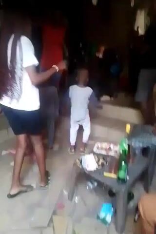 The video shows a group of people, including several children, gathered in what appears to be an outdoor or semi-outdoor setting. The ground is tiled. The children are dancing or moving around. There is a small plastic table with items on it, including what appear to be bottles, food on a plate, and cash. Some adults are watching. The scene seems to be a casual social gathering.