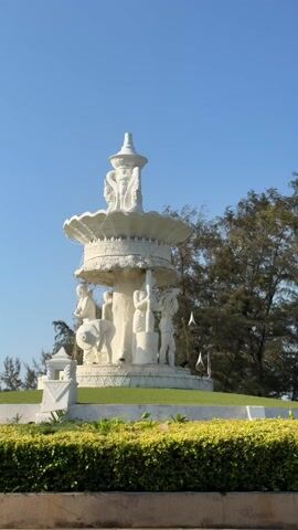 The video showcases a beautifully maintained public area featuring two prominent white statues under a clear, bright blue sky.

The central focus is a large, multi-tiered white fountain-like structure, intricately sculpted with ornate patterns and reliefs. The top tapers to a conical spire, while several bowl-shaped tiers descend, each adorned with detailed carvings. At the base of this structure, several human-like figures are depicted in white sculpture, appearing to be engaged in various activities. One figure is seen holding a lotus flower, another seems to be interacting with a wheeled object or drum, and another is shown standing with a long staff. These figures are surrounded by lush green grass on a raised mound.

In the foreground, vibrant, colorful flowers in shades of red, pink, orange, yellow, and white bloom abundantly, set against neatly trimmed green hedges.

As the camera pans slightly to the left, another impressive white statue comes into view. This statue, positioned on a tall pedestal, depicts a majestic bird, possibly a peacock or a mythical creature with elaborate tail feathers and a crest, facing away from the viewer. This bird statue is also made of white material and stands on a white architectural base with visible steps leading up to it.

The background consists of a line of tall, slender trees with dark green foliage. The scene is bathed in bright daylight, and the sky is a striking, cloudless blue. The overall impression is one of a serene and artistically rich outdoor space.