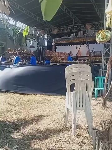 The video shows an outdoor stage set up for a performance. In the background, a large, stylized face is mounted above a screen, suggesting a traditional puppet show or theatrical performance. A group of people in blue shirts are seated on a raised platform behind a dark curtain, appearing to play musical instruments, possibly a gamelan orchestra. Several stacks of white plastic chairs are in the foreground, some empty and some with an audience member seated. The ground is covered in dry grass and leaves. The overall environment appears to be rural or an open field under a bright, sunny sky.