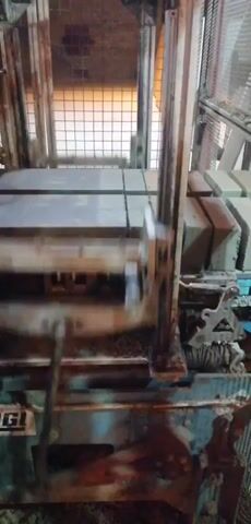 The video shows a close-up view of a brick-making machine in operation. The machine is made of metal and appears to be industrial-grade. There are several bricks visible on a conveyor belt to the right of the frame. The bricks are rectangular and appear to be made of a light-colored material. The machine has a large, heavy-duty arm that moves up and down, pressing the bricks. There are also several gears, levers, and other mechanical components visible. The environment appears to be a factory or workshop, with a wire mesh fence in the background. The lighting is dim, suggesting an indoor setting. The overall impression is one of an active industrial process.