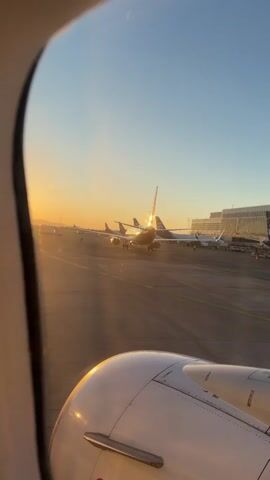 This is a POV video taken from inside an airplane looking out the window. The sun is setting, casting a warm, golden glow over the tarmac. Several other airplanes are parked on the ground, their tails illuminated by the setting sun. The camera pans slightly to the left, showing more of the parked aircraft and the airport terminal in the distance. The engine of the airplane the viewer is inside is visible in the foreground, partially obscuring the view. The overall mood is peaceful and contemplative, as one might feel while watching the sunset from a stationary aircraft.