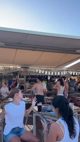 At 00:00, a young woman with long blonde hair, wearing a white t-shirt and denim shorts, is seated at a table. To her right, a woman with dark, curly hair, wearing a white tank top with a floral print, looks at her phone. A man with dark hair, wearing a black t-shirt and shorts, stands near a table in the background, holding a white towel. A glass of dark liquid sits on the table in front of the woman with curly hair. The scene is outdoors, with a large beige awning overhead. Bunting hangs along the edge of the awning. Numerous tables and chairs are visible, with other people in the background.

At 00:01, the woman with blonde hair is still seated at the table, looking to her left. The woman with curly hair is still looking at her phone. The man with the towel is still standing, but now slightly more to the left, and other people are visible around him. The background shows more people and tables.

At 00:02, the perspective shifts slightly. The woman with curly hair is still in the foreground, now with a second woman next to her who is wearing a white tank top with a floral pattern. The man with the towel is now further back, and a man in a white t-shirt and shorts is standing to his left. Several other people are scattered throughout the scene, some standing, some seated.

At 00:03, the view widens, showing more of the outdoor space. The man in the white t-shirt and shorts is now facing away from the camera, walking with another man who is also wearing a white t-shirt and shorts. In the background, a group of people are wearing blue and white striped shirts. More tables and chairs are visible, and some of the people appear to be dancing or celebrating.

At 00:04, the camera pans further to the right, revealing a stage area with musicians. People are gathered around the stage, some dancing. The overall atmosphere appears to be lively and festive, possibly at a beachside bar or restaurant, given the sandy ground. The sky is clear and blue.