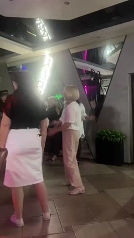 The video shows a group of people dancing in what appears to be a dimly lit restaurant or club. The perspective is from behind one of the dancers, a woman wearing a black top and a white skirt. Other people are visible dancing in the background, including a woman with blonde hair in a white top and beige pants, who is the focal point of the video. There are mirrors on the wall reflecting lights and more people. The floor is tiled, and there are plants in the background. The lighting is colorful, with pink and blue hues visible, along with a bright white light source hanging from the ceiling.