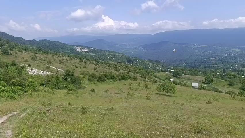 An aerial drone shot captures a panoramic view of a rural landscape on a clear day. The foreground is dominated by a grassy hill dotted with wildflowers and sparse bushes. In the mid-ground, the terrain slopes down into a valley dotted with scattered houses and trees. A small cultivated field with rows of plants is visible on the right side, enclosed by a rustic wooden fence. Further in the distance, a range of layered mountains forms the backdrop, with some cloud cover over their peaks. A white paraglider is suspended in the air over the valley, adding a touch of human activity to the otherwise serene natural environment. The overall impression is one of a peaceful, expansive countryside.