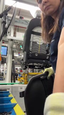 A woman wearing a dark blue t-shirt, black pants, and yellow gloves is shown in a factory setting. She is standing next to a car seat that is mounted on a yellow mechanical jig. The seat is black with white stitching and appears to be partially assembled. The woman is using her gloved hands to adjust a black fabric pouch on the back of the seat. In the background, another person is visible working at a conveyor belt, and to the left, a screen displays some data. The environment appears to be an assembly line with various mechanical components and industrial equipment. The woman is focused on her task, her head tilted slightly as she works on the seat.