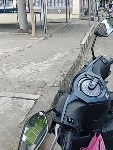 The video shows a street scene with a motorcycle in the foreground. The motorcycle is dark blue and has a digital dashboard. The background shows a paved area with a gray sidewalk and a building with a red roof. There are several people walking and riding motorcycles in the distance. The weather appears to be sunny.