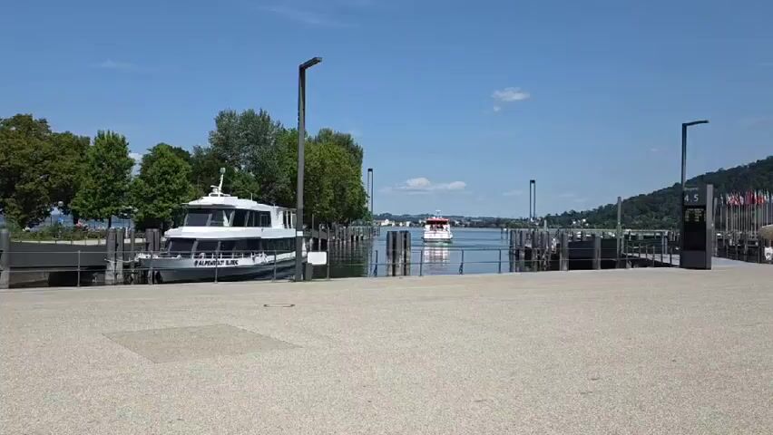 The video shows a sunny day at a harbor. A large white boat with a dark blue stripe is docked on the left side. The boat has a name printed on its side: "ALPENSTADT BLIES". Another smaller white boat with a red roof is moving away from the dock into the water. This boat has people visible on its deck. The harbor has several wooden posts and a metal railing. In the background, there are trees, buildings, and hills under a blue sky with some clouds. The ground in the foreground is a light-colored paved area.