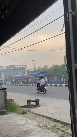 A man is riding a motorcycle on a street. He is wearing a white shirt and dark pants. There are other motorcycles and vehicles on the street. The sky is hazy and the sun is setting. There are buildings and shops on the side of the street. The scene appears to be in an urban or semi-urban area.