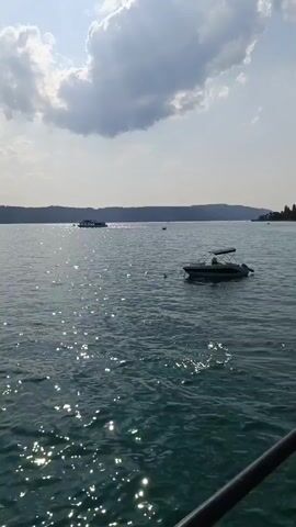 The video shows a wide shot of a large body of water, likely a lake or sea, under a partly cloudy sky. The water is a vibrant turquoise color and reflects the sunlight, creating a shimmering effect. In the foreground, a dark railing or structure is visible at the bottom edge of the frame, suggesting the viewpoint is from a dock or boat.

Several boats are present on the water. In the mid-ground, a small, white motorboat with a canopy is anchored or drifting. To the left, a larger, darker boat, possibly a ferry or tour boat, is moving away from the camera. In the distance, a hazy shoreline with trees and buildings is visible. The overall atmosphere is serene and suggests a pleasant day outdoors.