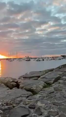 The video shows a sunset over the ocean. In the foreground, there are rocks. In the background, there are boats in the water and hills in the distance. The sky is cloudy and the sun is setting, casting a golden light over the scene. The video pans to the right, revealing a town on the shoreline. The buildings are lit by the setting sun.