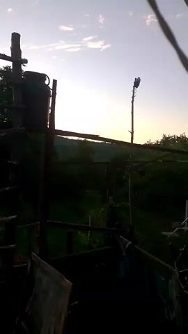 This video captures a slightly blurry, handheld view of an outdoor scene at dusk. The foreground is dominated by dark, shadowy structures and what appear to be frames or screens. In the mid-ground, there's a small, rustic-looking brick building with a corrugated metal roof. To the left of the building, several yellow objects and a white bucket are visible. Further back, a lush green landscape stretches out, with trees and foliage. Above the landscape, the sky is a soft gradient of blue and yellow, indicative of sunset, with a few scattered clouds. A tall, thin pole with some kind of equipment on top stands prominently in the middle distance. In the far left, a dark barrel or tank is perched atop a wooden structure. The overall impression is of a rural or semi-rural setting, possibly a backyard or farm area, during the transition from day to night.
