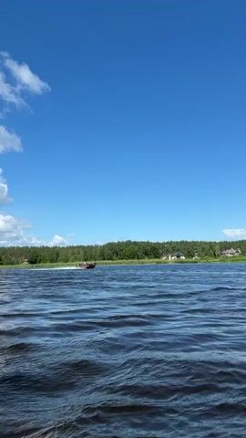 A motorboat is seen speeding across a large body of water, leaving a white wake behind it. The boat is red with white and black lettering that reads "WINEDIRECT". Two people are visible in the boat, one at the helm and another seated behind. The background features a lush green shoreline with trees and several houses. The sky is a clear, vibrant blue with a few scattered white clouds. The water appears choppy with small waves. The overall scene suggests a sunny day on a lake or river.