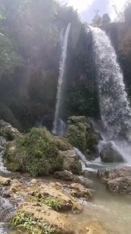 The video shows a waterfall with two main streams of water cascading down a rocky cliff face. Lush green vegetation covers the surrounding cliffs. In the foreground, there is a pool of clear, shallow water with rocks scattered throughout. Towards the right side of the frame, a woman in a light-colored bikini stands on a large rock. In the background, other people are visible near the water. The scene appears to be a natural, outdoor environment.