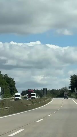 The video shows a perspective from inside a vehicle traveling on a highway. The road curves to the right, with two lanes visible. On the left side of the road, there's a grassy shoulder with a wooden fence and vegetation. Several vehicles are seen on the highway, including white vans and red trucks, and a black car in the distance. In the background, there are trees and a cloudy sky. A large, dark rectangular sign with a metal frame is visible on the left side of the road, facing away from the camera. The video appears to be shot during daylight.