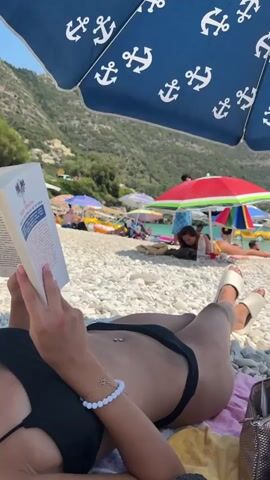 A low-angle medium shot shows a person lying on a beach towel reading a book. The person is wearing a black bikini and white sandals. They are lying on their back with their legs extended and their head is out of frame. The person's hands are visible holding the book open. A beaded bracelet is on their left wrist. The book is titled "Le Monde" and has a French flag on its cover. In the background, a blue umbrella with white anchor patterns is overhead. Beyond the umbrella, a crowded beach is visible with several people sunbathing and swimming. There are colorful beach umbrellas, lounge chairs, and people scattered across the pebble beach. The ocean is a clear turquoise color, and a green, tree-covered hillside rises in the background.