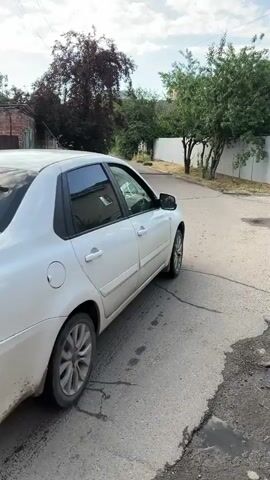 This is a vertical video clip taken in Krasnodar, Russia. The scene shows the rear of a white Datsun sedan parked on a paved road. The car has a license plate that reads "M569XP 123" and the text "RUS" is visible below it. The car appears to be stationary.

The setting is an outdoor, possibly residential or industrial, area with a brick wall and a tall fence visible on the left and right sides of the frame, respectively. Lush green trees and foliage are present behind the fence on the right and also to the left of the road, suggesting it is summertime. The sky is partly cloudy, indicating it is daytime. The road surface is cracked and worn, with some puddles of water present, suggesting recent rain or damp conditions. The overall atmosphere of the video is neutral, focusing on documenting the car and its surroundings. There are no people visible in the clip.