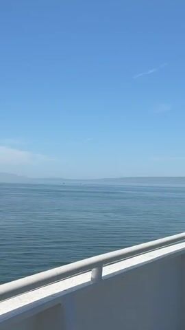 A clear blue sky is visible above a calm blue ocean. A distant landmass is faintly visible on the horizon. Two small boats are moving across the water. The foreground shows a white railing and deck, suggesting the video is filmed from a boat.
