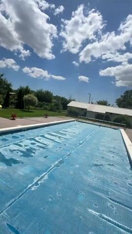This video pans across an outdoor scene featuring a swimming pool covered with a blue solar blanket. The pool area is surrounded by a tiled patio and lush green grass. In the background, there are buildings, trees, and a bright blue sky with fluffy white clouds, suggesting a sunny day. The overall impression is one of a well-maintained recreational area, likely a backyard or a communal space, in a suburban or rural setting. The time of day appears to be daytime, given the bright sunlight and clear sky. There is no visible text in the video.
