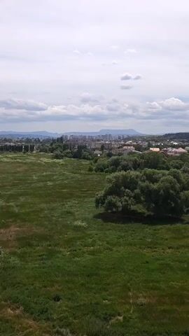 A panoramic view of a city is captured from a slightly elevated position. In the foreground, a vast expanse of lush green grass stretches across the frame, with a few scattered trees dotting the landscape. The middle ground is dominated by a dense cluster of trees, varying in shades of green, that create a natural barrier between the open field and the urban sprawl. Beyond the trees, the cityscape unfolds, characterized by a mix of buildings. Several tall, modern apartment buildings, mostly white and grey, punctuate the skyline, interspersed with lower-rise residential structures. The architecture suggests a mix of Soviet-era and contemporary designs. The background features rolling hills and mountains under a partly cloudy sky. Construction cranes are visible in the distance, indicating ongoing development within the city. The overall impression is of a city blending with its natural surroundings, with a clear delineation between green spaces and urban development.