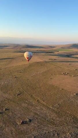 The video captures a serene hot air balloon flight over a vast, open landscape, likely in Yeşilhisar, Turkey, given the provided context. The time of day appears to be early morning, possibly sunrise, with a clear blue sky transitioning to a soft yellow and orange glow near the horizon.

Multiple hot air balloons are visible in the scene. In the opening shot, a white balloon with "Turkiye" written in red text is prominent, floating relatively low over dry, brown fields with patches of green. As the camera pans, more balloons come into view, including yellow ones, ascending higher into the sky. The landscape consists of rolling hills, plains, and distant mountains, with some areas appearing cultivated and others more arid. A body of water, possibly a lake or reservoir, is also seen nestled between hills, reflecting the morning light. The overall atmosphere is calm and expansive, highlighting the unique experience of a hot air balloon ride over such a picturesque terrain.