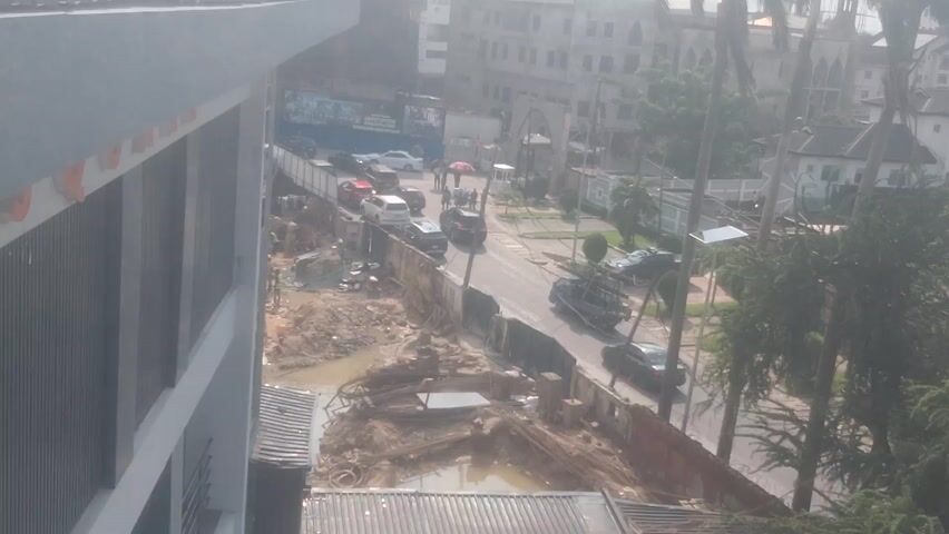 This is an overhead view of a street construction site. A building is visible on the left side of the frame. The street is lined with traffic, including cars, a truck, and what appears to be a construction vehicle. The construction site itself is characterized by piles of dirt, mud, and rebar. Trees and other buildings are visible in the background, suggesting an urban environment. The lighting indicates it is daytime, possibly overcast or hazy.