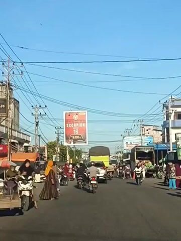The video captures a busy street scene during daylight hours. The sky is a clear blue with scattered utility wires crisscrossing overhead. A prominent billboard advertising "SCORPION 16" is suspended across the street. The road is filled with various vehicles, including numerous motorcycles, cars, and a truck. People are seen riding on the motorcycles and walking along the sides of the road. Some individuals are dressed in traditional clothing, including headscarves and long dresses. On the left side of the road, there are market stalls with some produce displayed. The overall atmosphere suggests a lively urban or semi-urban environment.