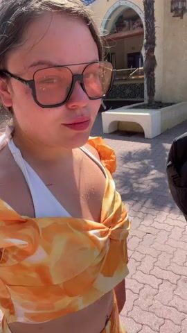 This is a first-person perspective video, likely captured on a smartphone, showing a young woman with sunglasses and a white and orange swimsuit. She appears to be at an outdoor amusement park or a similar recreational area. She is smiling and looking around, suggesting she is enjoying herself.

In the background, a man with sunglasses and a backpack is visible, holding a drink. He is walking past a food stall with a sign that reads "PATATE FRIES." Further in the background, a roller coaster structure is visible against a bright blue sky, indicating a sunny day. The architecture of the buildings in the background has an exotic, possibly Middle Eastern or North African influence, with arched doorways and decorative elements. Palm trees and other greenery are present, suggesting a warm climate. The ground is paved with interlocking bricks. The overall atmosphere is casual and festive, consistent with a day out at an amusement park. The time of day appears to be daytime, given the strong sunlight and shadows.