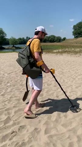 A man with a beard, wearing a yellow t-shirt, white shorts, a white baseball cap, and beige sandals, walks across a sandy beach. He has a large green backpack on his back, with a shovel attached to the side of the backpack. He is holding a yellow and black metal detector and appears to be sweeping it across the sand. The sun is shining, and the sky is clear blue. In the background, there is a body of water with trees along the far bank. The sand is marked with footprints and shadows.