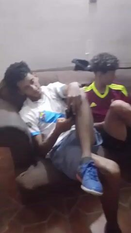 Three young men are sitting on a brown couch indoors. The man on the left is wearing a white and blue soccer jersey, gray shorts, and blue shoes. He is looking down at a phone in his hands. The man in the middle has curly hair and is wearing a maroon and gold soccer jersey with black shorts. The man on the right is wearing a red shirt with a black and white stripe across the chest and black shorts. He is smiling and looking at the person holding the camera. The walls are light gray. There is a glass on a shelf in the background. The floor is tiled. The camera pans slightly from left to right.