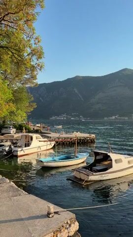 The video captures a serene waterfront scene in Dobrota, Montenegro, likely during the late afternoon or early evening, given the warm, golden light casting a gentle glow. The calm, dark water of the bay reflects the clear, bright blue sky, with rolling green mountains forming a dramatic backdrop.

Several small boats are moored along a stone quay in the foreground. Two white motorboats, one with a covered section, are prominent. Between them, a smaller, light blue rowboat bobs gently. A larger white boat, docked further back, has its stern facing the viewer. The name "Stefani" is visible on the side of one of the motorboats.

Along the shore to the left, a tree with lush green foliage casts dappled shadows. A few people can be seen walking or standing near the water's edge, enjoying the tranquil atmosphere. A small stone pier extends into the water, with a few more boats docked alongside it. In the distance, a line of buildings suggests a settlement nestled along the bay's edge, with a faint trail of smoke rising from a chimney. A single, distant boat can be seen moving across the water.

The overall mood is peaceful and idyllic, conveying a sense of quiet relaxation and the natural beauty of the Montenegrin coast. The scene evokes a feeling of calm and invites contemplation of a simple, beautiful day.
