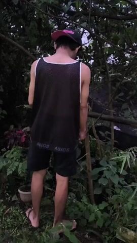 A man is standing outdoors in a densely vegetated area. He is wearing a black and white mesh tank top with the word "ASICS" repeated around the bottom, black shorts, and flip-flops. He has a black baseball cap with a pink brim turned backward on his head. He appears to be looking at or touching a small tree or branch. The environment is green and overgrown, with various plants and small trees visible. The lighting suggests it is either dusk or a heavily shaded area.