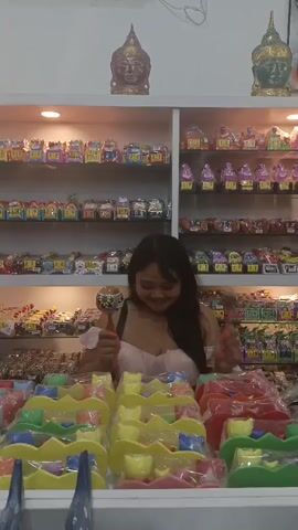 A woman in a pink dress stands behind a counter filled with colorful toys and trinkets, holding two small maracas. She shakes them rhythmically, smiling at the camera. The shelves behind her are stocked with similar items, and two Buddha head statues are displayed on the top shelf. The scene appears to be a small shop, possibly a souvenir shop.