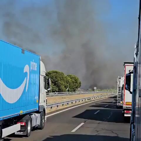 This video shows a wildfire from the perspective of a vehicle traveling on a highway. Thick plumes of dark gray smoke billow into the sky from a wooded area to the left of the highway. The smoke appears to be spreading and obscuring the sky. A large blue truck with "prime" written on its side is in the foreground on the left, and the back of a white truck is visible on the right. The highway has a metal guardrail separating it from the dry, grassy shoulder and the burning vegetation. The sky is clear and blue above the smoke. There are no visible people in the immediate foreground, and the focus is on the road and the approaching smoke from the wildfire.