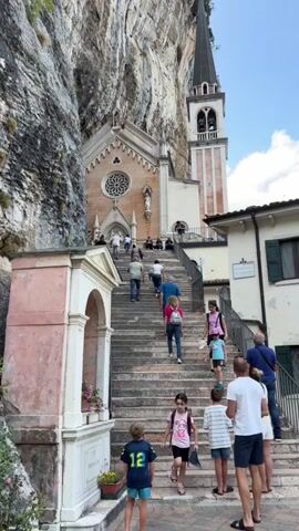 A group of people are walking up a long stone staircase leading to a church built into the side of a cliff. The church has a bell tower and a rose window. Some people are young children, while others appear to be adults. The weather is sunny and the sky is blue.