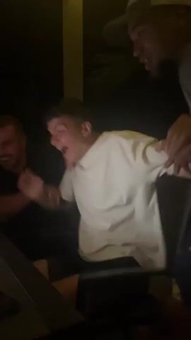 A group of three young men are gathered around a table outside at night, illuminated by string lights overhead. One man in a white shirt is seated at the table, looking excitedly at a laptop screen.  He is visibly surprised and reacts emphatically, holding his hands up to his face. Another man, wearing a black shirt, also seated, reacts with his hand covering his mouth in apparent astonishment. The third man, standing behind the seated man in white, puts his arms around the seated man in white. The laptop screen shows an online slot machine game with colorful graphics. In the background, there is a bottle of water and a package of what appears to be snacks. The overall atmosphere is one of excitement and surprise.