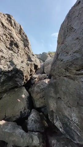 A person is climbing over large rocks on a sunny day. The person is wearing dark shorts and appears to be bare-chested. The rocks are rough and varied in size. The sky is clear blue with some white clouds and the sun is visible. There are some trees and structures in the background.